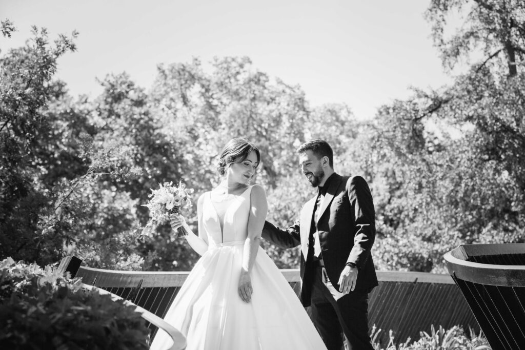 Le marié regarde le dos de la robe de la mariée. Il découvre avec émotion les détails. La mariée souriante apprécie le moment sur un pont avec de la verdure