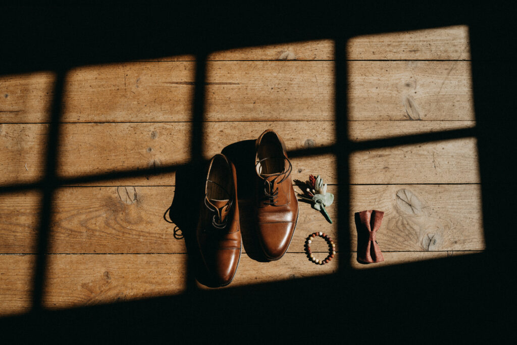 Photo de détail des accessoires du marié : chaussures marrons, bracelet, boutonnière en fleurs, noeud papillon