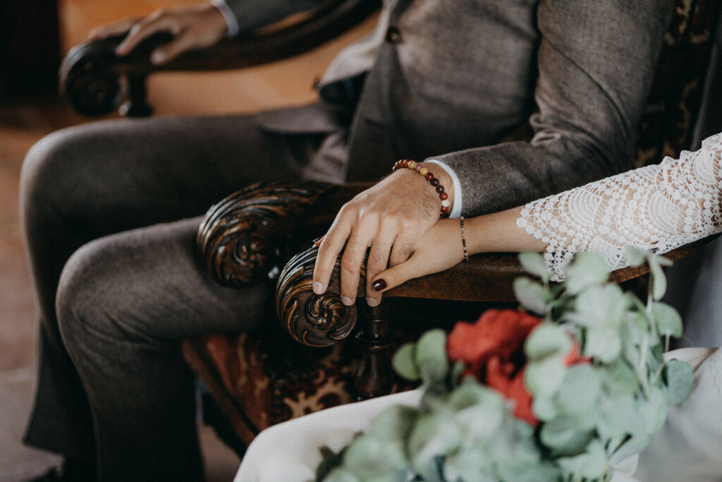 Couple de mariés se tenant mains dans la main pendant qu'ils écoutent leur cérémonie laïque assis sur des fauteuils le bouquet à la main 