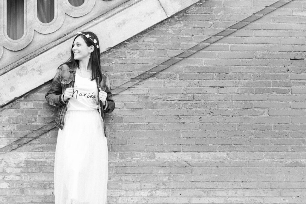 EVG, EVJF la mariée adossée à un mur en tutu et tee shirt blanc veste en jean couronne de fleur souriante. Photo en noir et blanc 