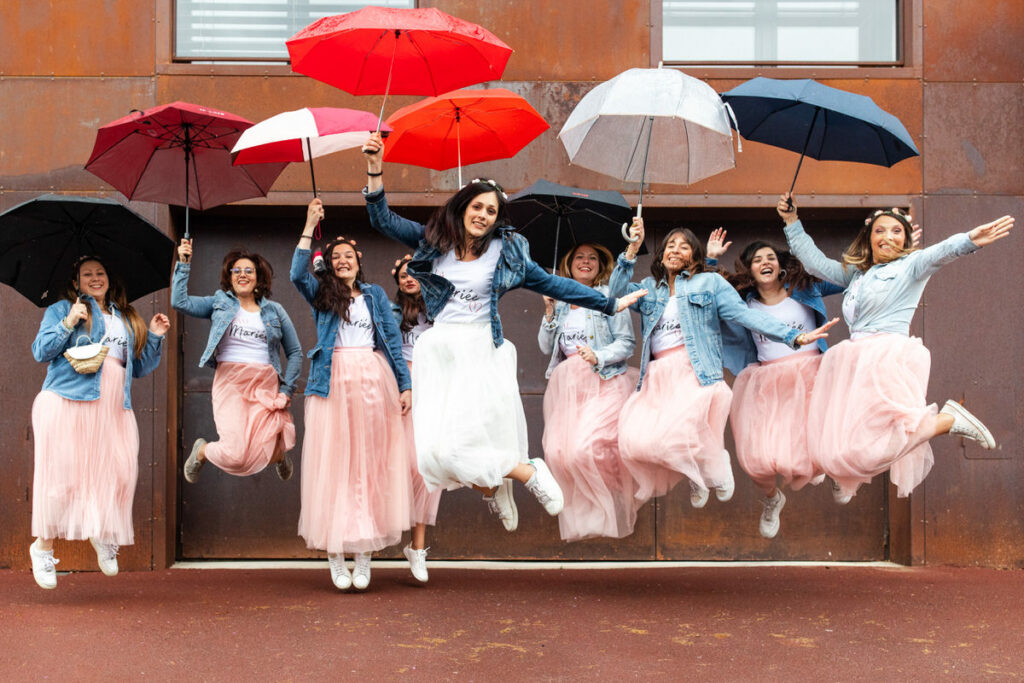 EVG, EVJF la mariée et ses témoins demoiselles d'honneur sautent avec des parapluies. vêtues de tutu rose de tee shirt blanc et veste en jean 